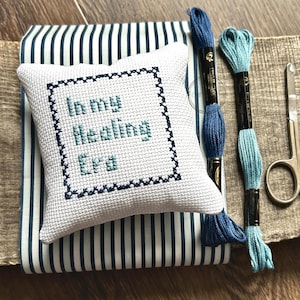 The finished pillow, made from this cross stitch pattern, is sitting on a board with all of the supplies.