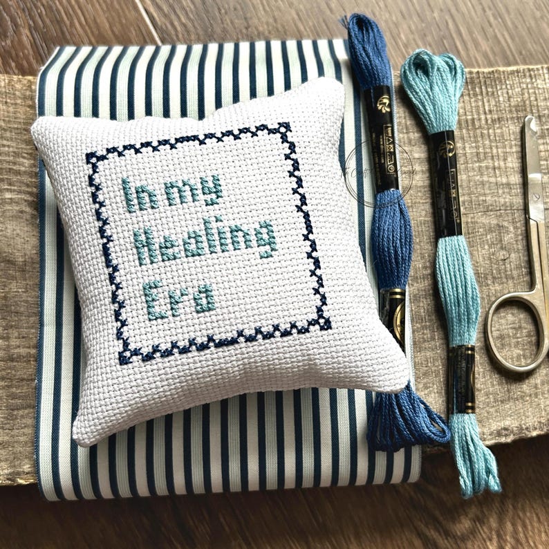 The finished pillow, made from this cross stitch pattern, is sitting on a board with all of the supplies.