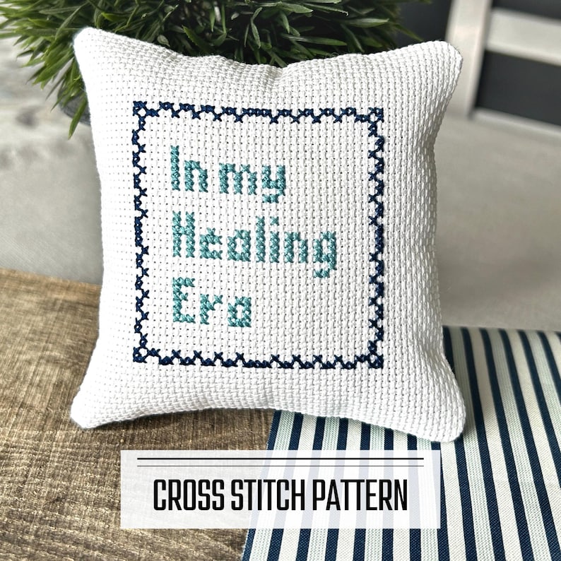 A DIY cross stitch pattern for a mini pillow that can be used as a pin cushion. The finished pillow is sitting on a wooden board.