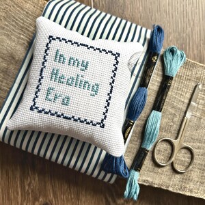 The finished pillow, made from this cross stitch pattern, is sitting on a board with all of the supplies.