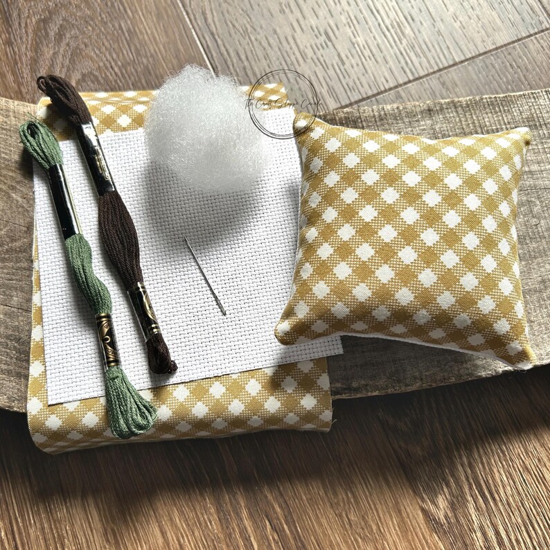 The supplies needed to make this cross stitch mini pillow are sitting on a wooden board.