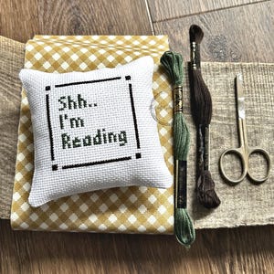 The finished pillow, made from this cross stitch pattern, is sitting on a board with all of the supplies.