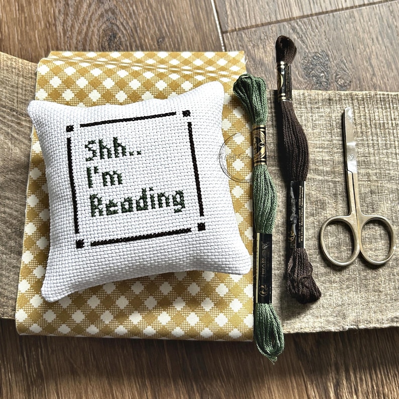 The finished pillow, made from this cross stitch pattern, is sitting on a board with all of the supplies.