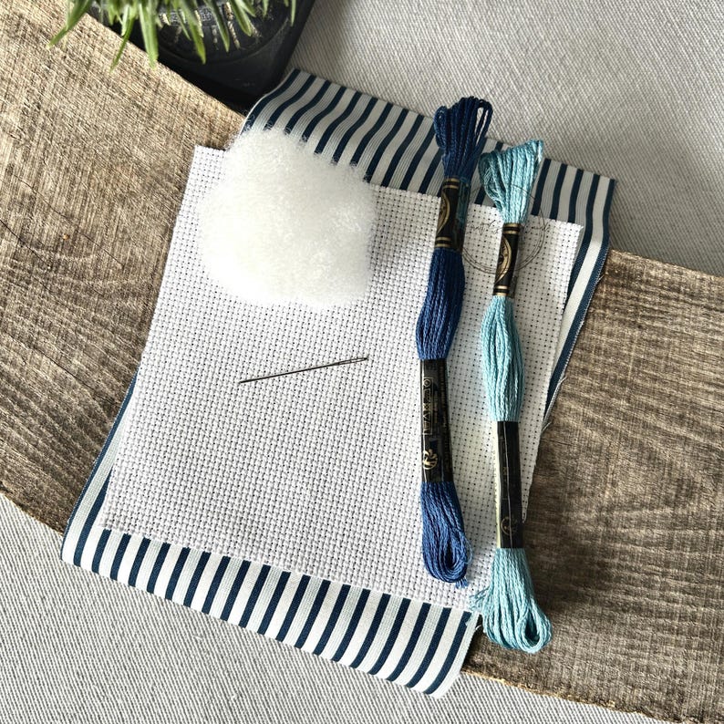 The supplies needed to make this cross stitch mini pillow are sitting on a wooden board.