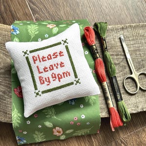 The finished pillow, made from this cross stitch pattern, is sitting on a board with all of the supplies.