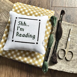 The finished pillow, made from this cross stitch pattern, is sitting on a board with all of the supplies.