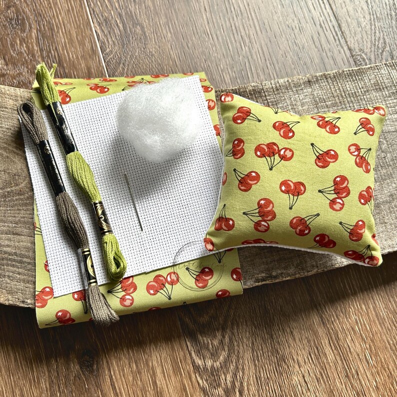 The supplies needed to make this cross stitch mini pillow are sitting on a wooden board.