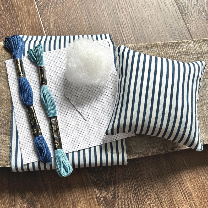 The supplies needed to make this cross stitch mini pillow are sitting on a wooden board.