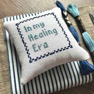 The finished pillow, made from this cross stitch pattern, is sitting on a wooden board.
