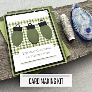 May include: A handmade Christmas card with olive green bulb ornaments and the text "You Make Christmas A Little Brighter." The card is layered with black and green accents, and a spool of twine and adhesive tape are nearby. The words "CARD MAKING KIT" are at the bottom.