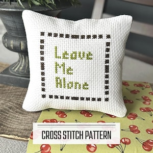 A DIY cross stitch pattern for a mini pillow that can be used as a pin cushion. The finished pillow is sitting on a wooden board.