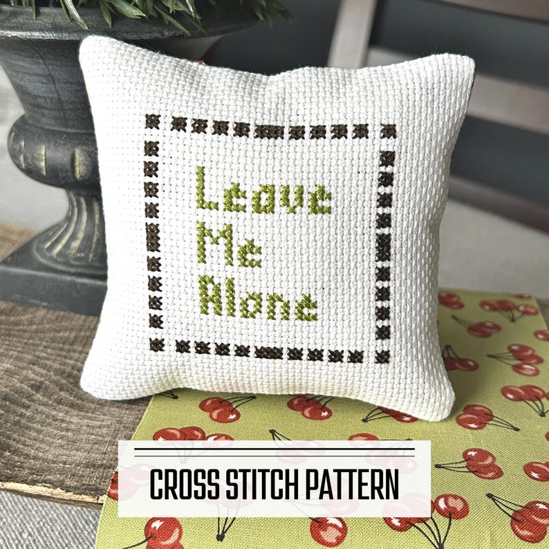 A DIY cross stitch pattern for a mini pillow that can be used as a pin cushion. The finished pillow is sitting on a wooden board.