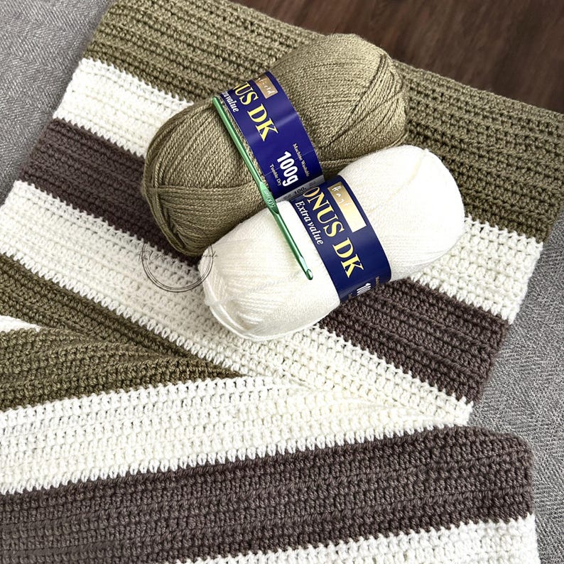 The finished baby blanket, made from this crochet pattern, is draped over a couch with 2 balls of yarn and a crochet hook.