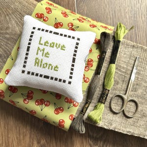 The finished pillow, made from this cross stitch pattern, is sitting on a board with all of the supplies.