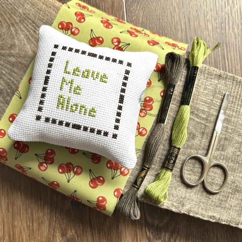 The finished pillow, made from this cross stitch pattern, is sitting on a board with all of the supplies.