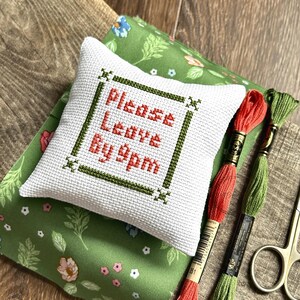 The finished pillow, made from this cross stitch pattern, is sitting on a board with all of the supplies needed.