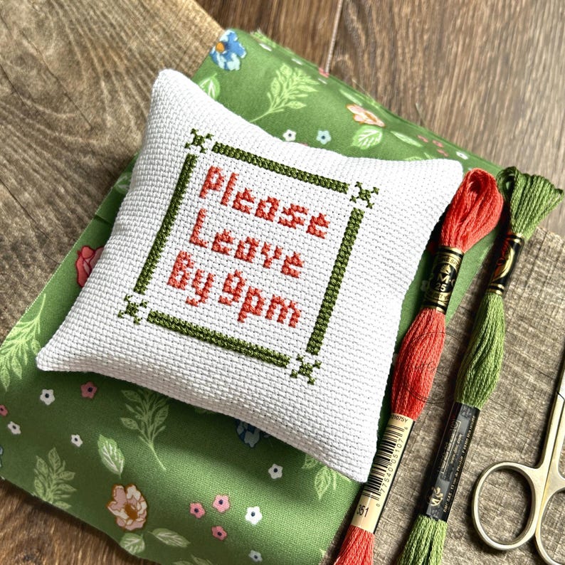 The finished pillow, made from this cross stitch pattern, is sitting on a board with all of the supplies needed.
