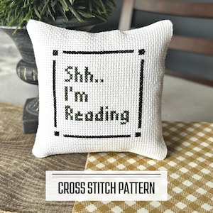 A DIY cross stitch pattern for a mini pillow that can be used as a pin cushion. The finished pillow is sitting on a wooden board.