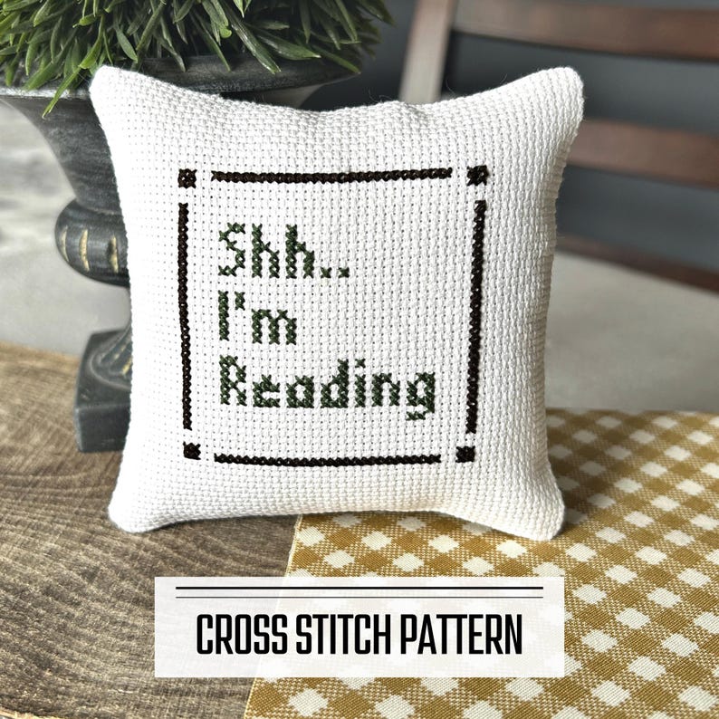 A DIY cross stitch pattern for a mini pillow that can be used as a pin cushion. The finished pillow is sitting on a wooden board.