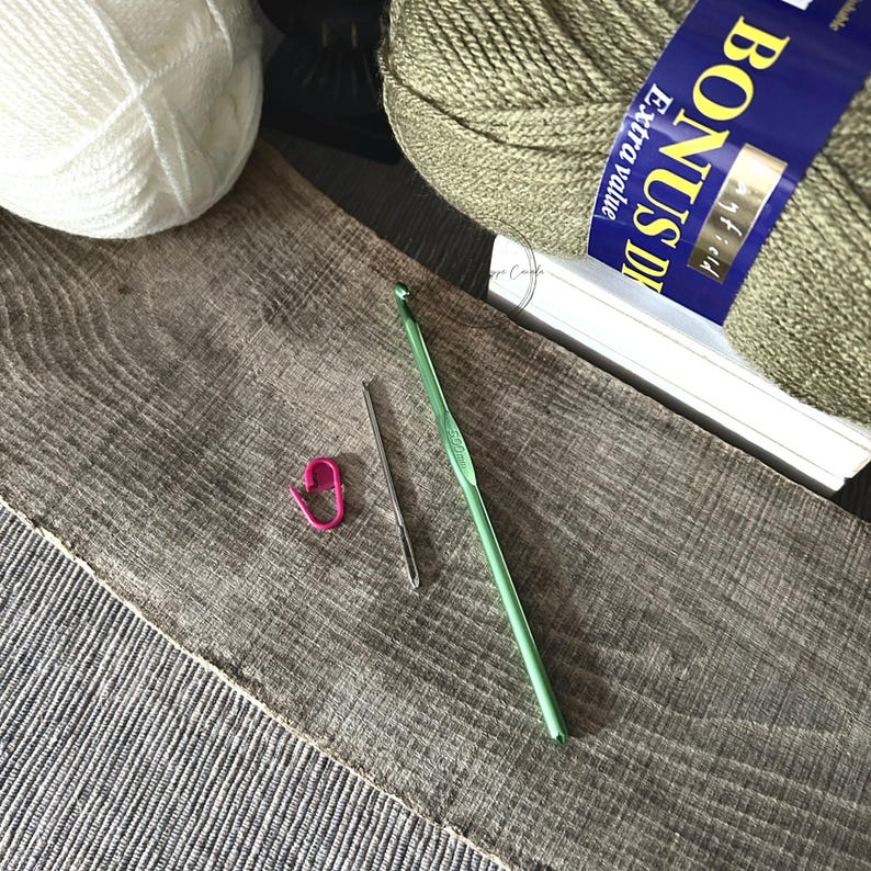 The tools needed to put the baby blanket together are lying on a wooden board. There are 2 balls of yarn, a crochet hook, tapestry needle and stitch marker.