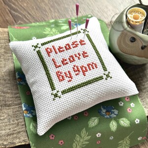 The finished pillow, made from this cross stitch pattern, is sitting on a wooden board with pins sticking out.