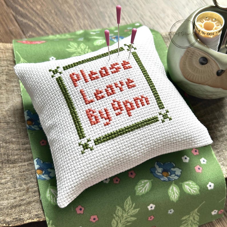 The finished pillow, made from this cross stitch pattern, is sitting on a wooden board with pins sticking out.