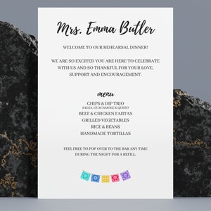 May include: A white card with black text announcing a rehearsal dinner for Mrs. Emma Butler. The card includes a menu with items such as chips and dip, beef and chicken fajitas, grilled vegetables, rice and beans, and handmade tortillas. The card is decorated with colourful paper flags.