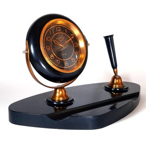 May include: A black desk clock with a gold face and Roman numerals. The clock is mounted on a black base with a pen holder. The clock is set to 11:20.