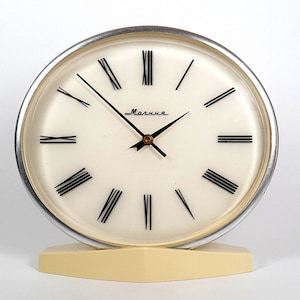 May include: A white analog clock with Roman numeral markings and a silver rim. The clock is on a cream-colored base and reads "Маякша".