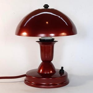May include: A red metal table lamp with a mushroom-shaped shade. The lamp has a black cord and a black on/off switch.