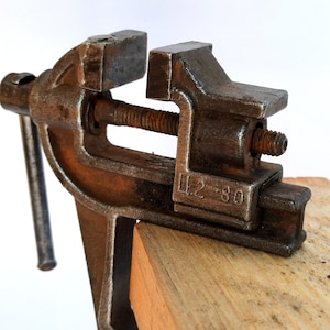 Peut inclure: Un étau en métal rouillé, fixé sur un bloc de bois. L'étau, de couleur marron foncé, présente un mécanisme à vis pour serrer et desserrer. Le texte "U.2-80" est visible sur le côté, indiquant son ancienneté.