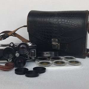 Puede incluir: Una cámara vintage negra y plateada con una funda de cuero y varios filtros para lentes. La cámara tiene una correa de cuero negra y la funda tiene un patrón de cocodrilo.