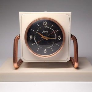 May include: A white and copper clock with a black face and gold hands. The clock is on a white base.