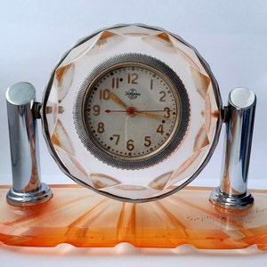 May include: A vintage clock with a clear, faceted glass dome and a white face with black numerals. The clock is mounted on a chrome stand with an orange base. The base has the inscription "Secunda, Switzerland".
