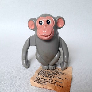 May include: A gray plastic monkey toy with large black eyes and a pink face. The toy is standing on a piece of paper with text in Russian that reads: "Игрушка для детей дошкольного и младшего школьного возраста" which translates to "Toy for preschool and primary school age children".