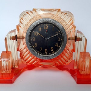 May include: An Art Deco style clock with a black face and gold numerals. The clock is encased in a translucent orange-red plastic with a fan-like design. The clock's face is surrounded by a silver-colored metal ring.