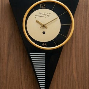 May include: A black and gold wall clock in a triangular shape. The clock face is cream with black numerals and hands. The clock's frame is black with a gold circular border. The clock has a unique, modern design, suitable for any room.