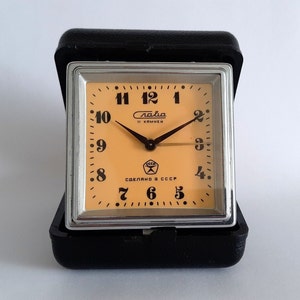 May include: A black alarm clock with a silver face and orange hands. The clock face has black numbers and the text "Слава 11 камней" and "СДЕЛАНО В СССР".