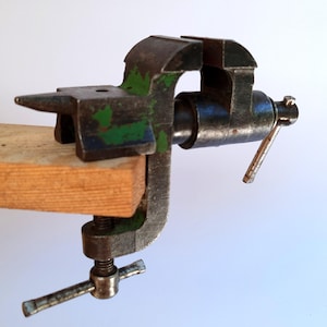Puede incluir: Una prensa de metal vintage, pintada de verde y azul, sujeta a un trozo de madera. La prensa tiene un mango y un mecanismo de tornillo para apretar. La herramienta está diseñada para sujetar objetos de forma segura, mide unos 15 cm.