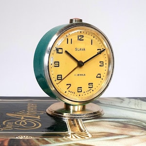 Può includere: Un orologio da sveglia vintage verde e oro con un quadrante giallo. Il quadrante dell'orologio ha numeri e lancette neri, e dice "Slavia 11 Jewels" in lettere nere. L'orologio è appoggiato su un libro.