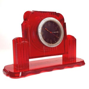 May include: A red plastic clock with a black face and silver hands. The clock is on a red plastic base with a scalloped edge.