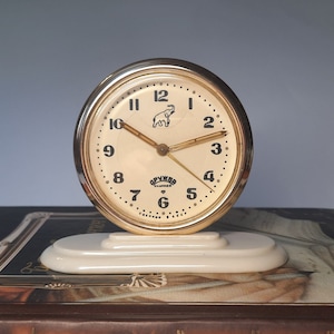 May include: A gold-coloured alarm clock with a white face and black numbers. The clock has a small elephant illustration on the face and the text "СССР" on the bottom. The clock is sitting on a white base.