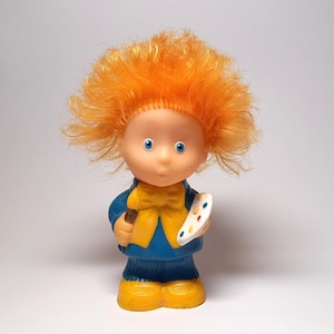 Op de afbeelding: Een pop met rood haar, blauwe ogen en een blauwe en gele outfit houdt een kwast en een palet.