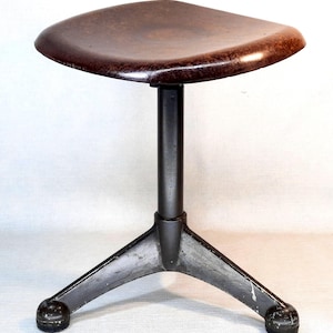 May include: A vintage, adjustable height stool with a dark brown, square seat. The seat is supported by a black, cylindrical post and a three-legged base. The base has rounded feet. The stool has a classic, industrial design.