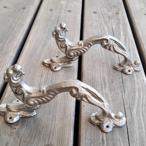 Peut inclure: Deux poignées de porte en métal argenté en forme de dragons stylisés avec des détails complexes. Les poignées sont présentées sur un fond en bois.