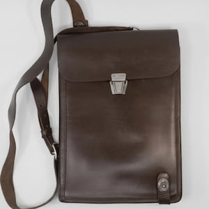May include: A brown leather messenger bag with a rectangular shape and a flap closure. The bag features a long, adjustable shoulder strap and a silver-colored clasp. The bag is approximately 12 inches tall and 10 inches wide.
