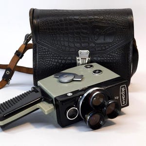 Puede incluir: Cámara de cine Ekran-4 vintage en negro y gris, con múltiples lentes y un asa. Acompañada de una funda de transporte de cuero negro con correa para el hombro. La funda y la cámara tienen texto. Una cámara clásica.