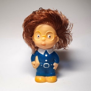 Op de afbeelding: Een kleine plastic pop met bruin haar, die een blauw en wit uniform draagt met een geel boek in haar hand.