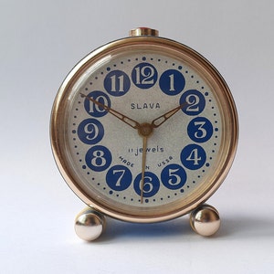 Könnte beinhalten: Ein goldfarbener Wecker mit weißem Zifferblatt und blauen Zahlen. Die Uhr hat einen roten Mittelknopf und trägt die Aufschrift "Slavia 11 Jewels Made in USSR" auf dem Zifferblatt.