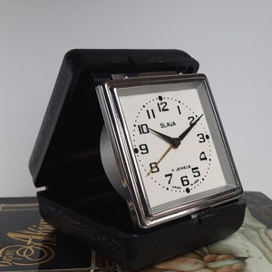 May include: A black and silver alarm clock with a white face and black hands. The clock face reads "Slavia" and "11 Jewels Made in USSR".